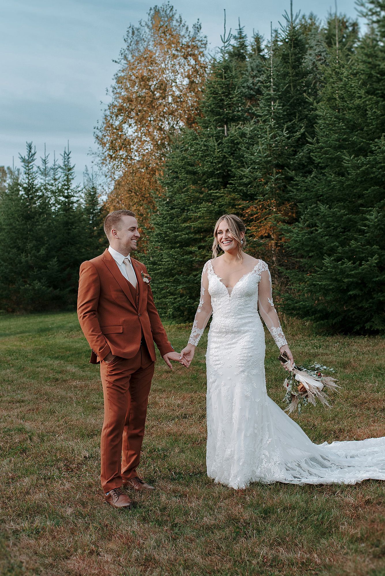 A couple in wedding attire hold hands while standing in a field with evergreen trees and autumn foliage in the background.