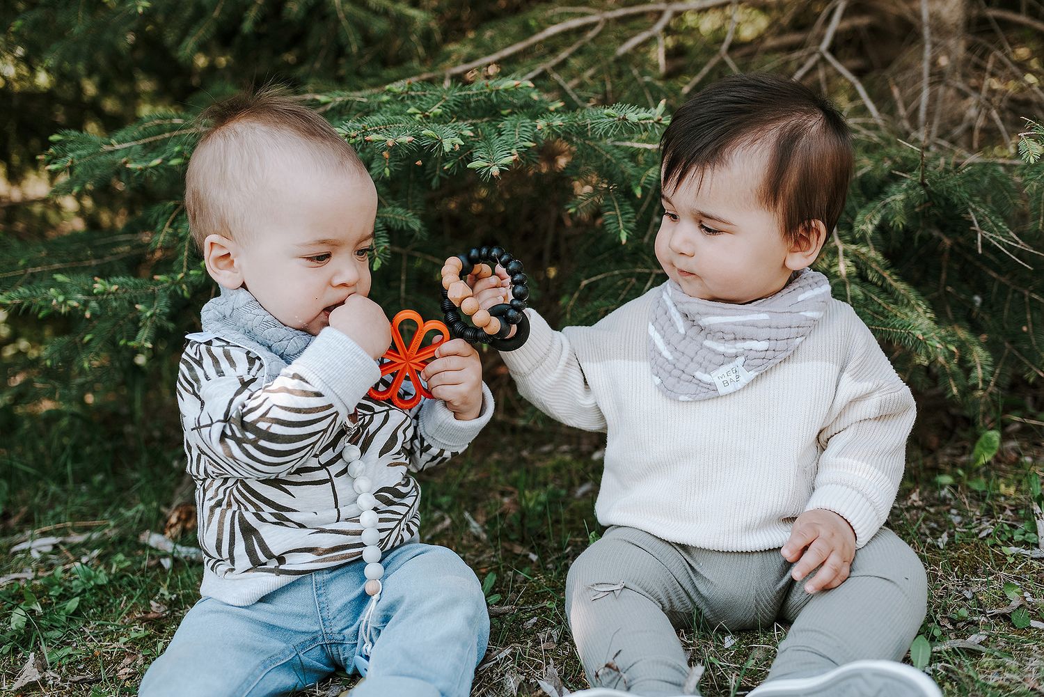Two babies in coordinating outfits sitting together on grass near evergreen trees.