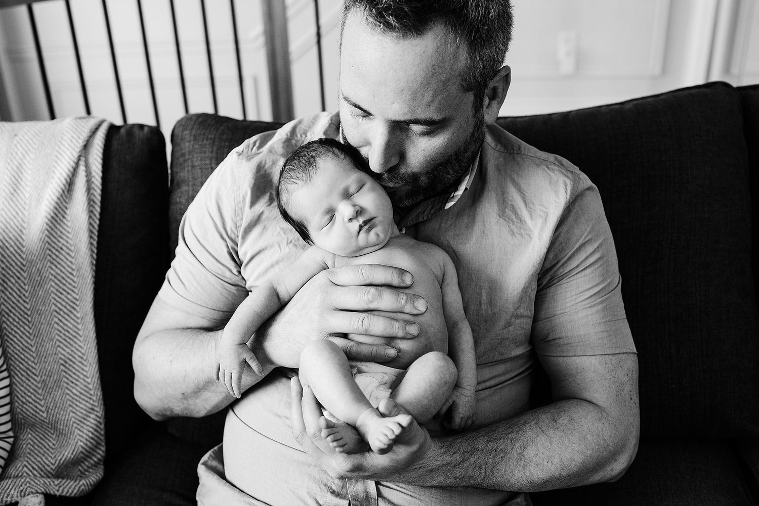 A black and white image of a parent holding and kissing a newborn baby on a dark couch.