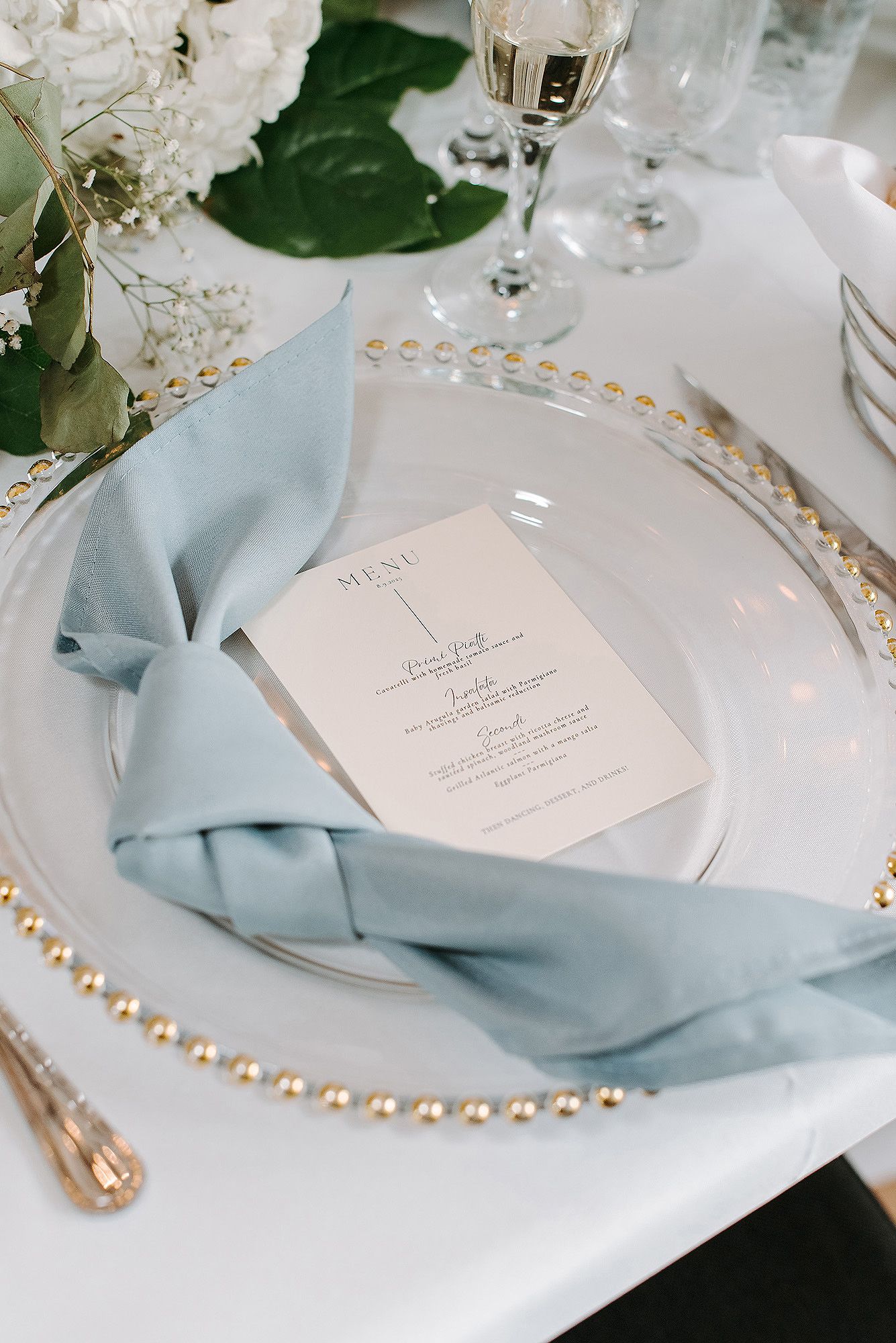 Elegant place setting with light blue napkin, beaded charger plate, and white menu card on formal dining table.