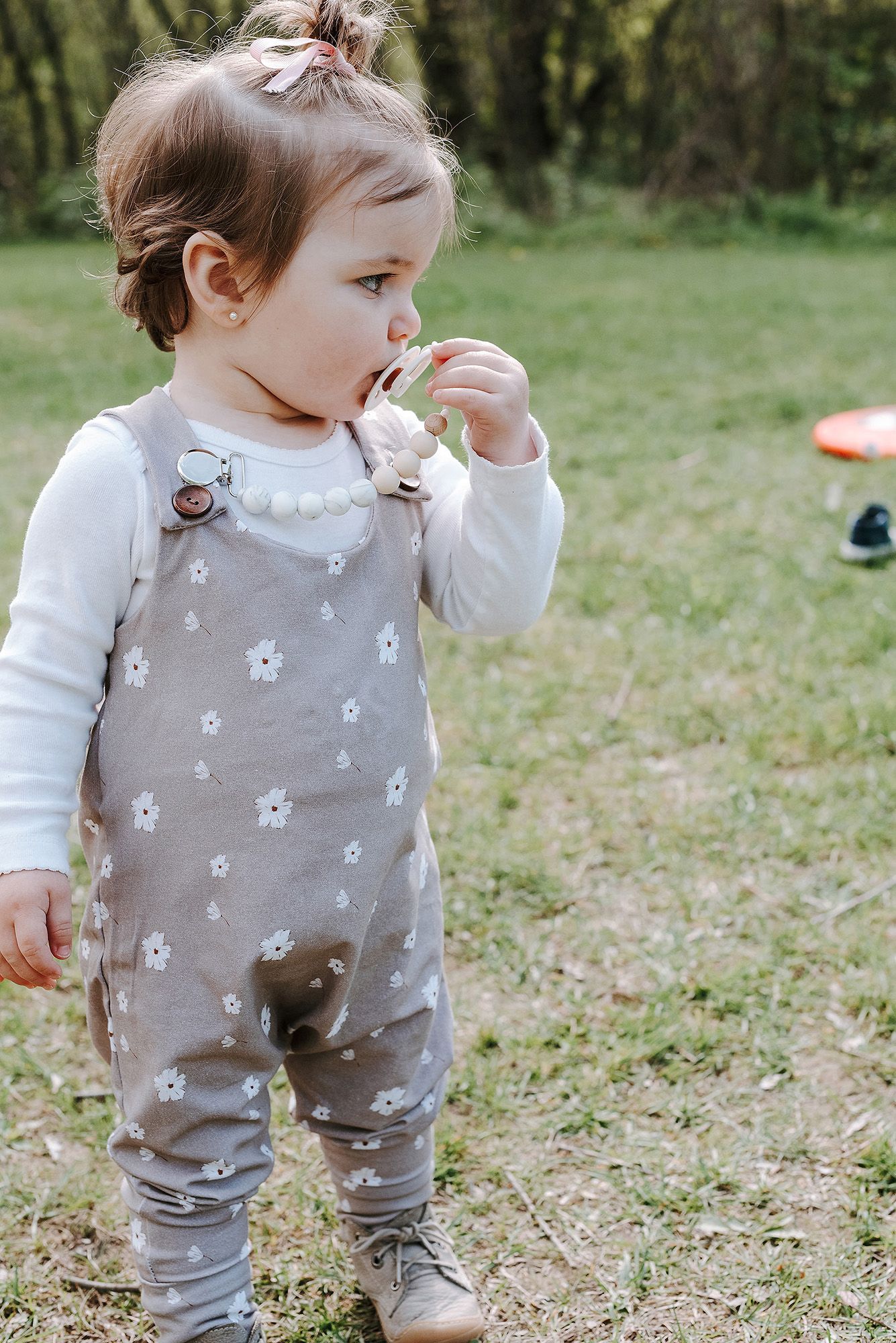 A toddler wearing gray floral overalls and white shirt stands in a grassy outdoor setting.