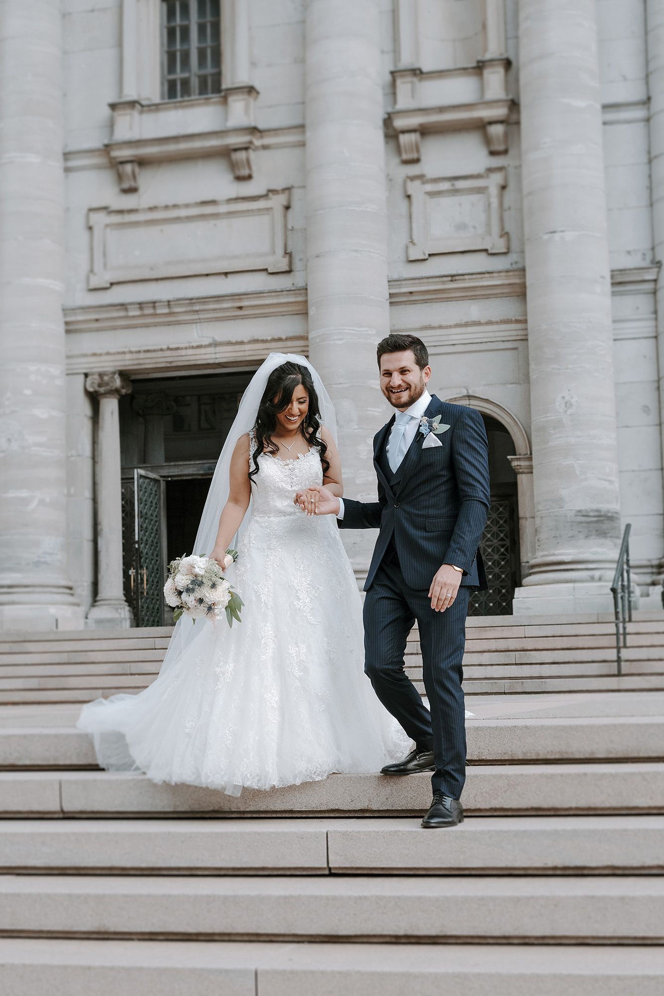 A bride and groom stand on grand stone steps outside a classical building with white columns on their wedding day.