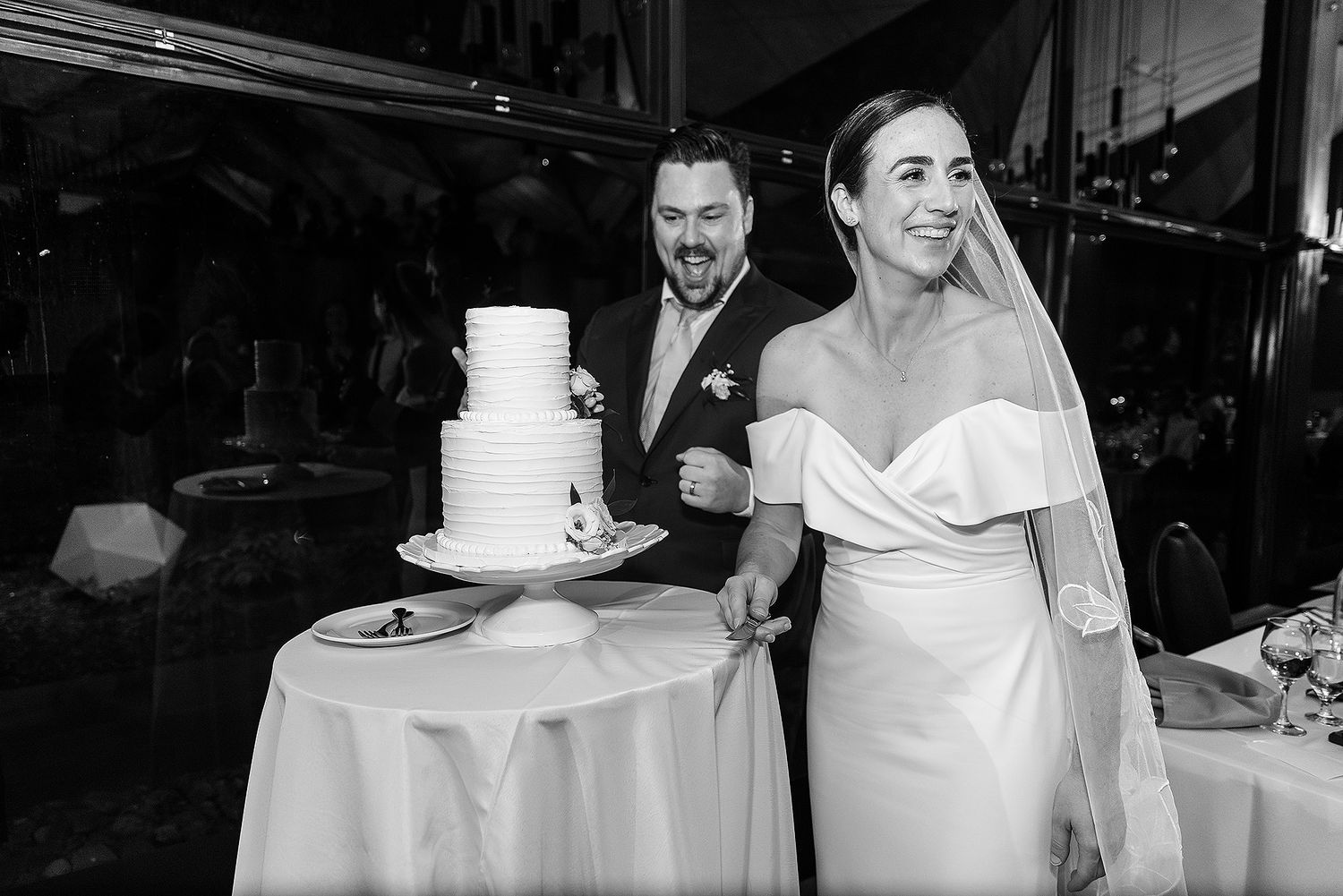 A black and white wedding photo showing a couple standing next to their elegant three-tiered white cake.