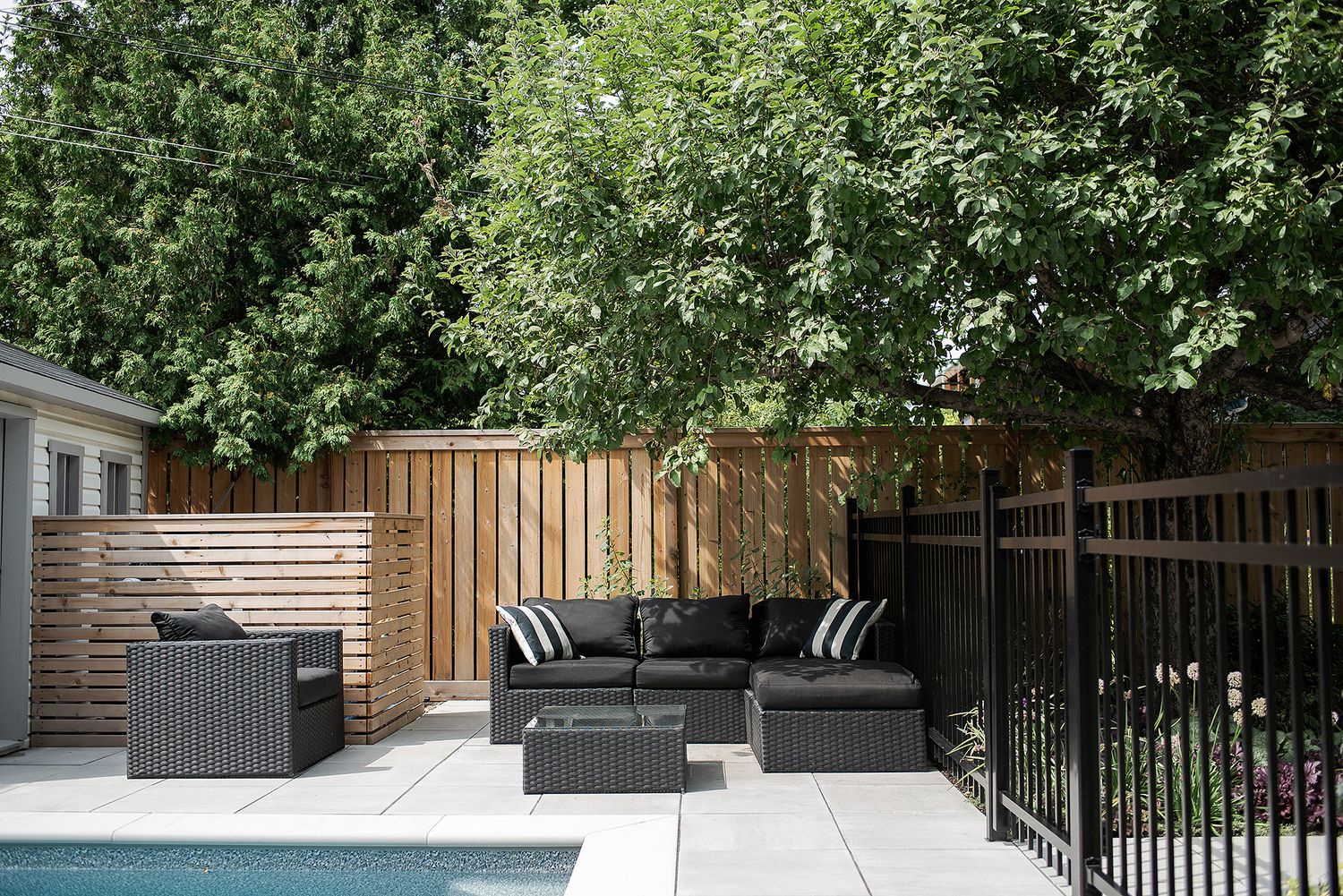 Modern outdoor patio area with wooden privacy fence, gray cushioned seating, and lush green trees overhead.