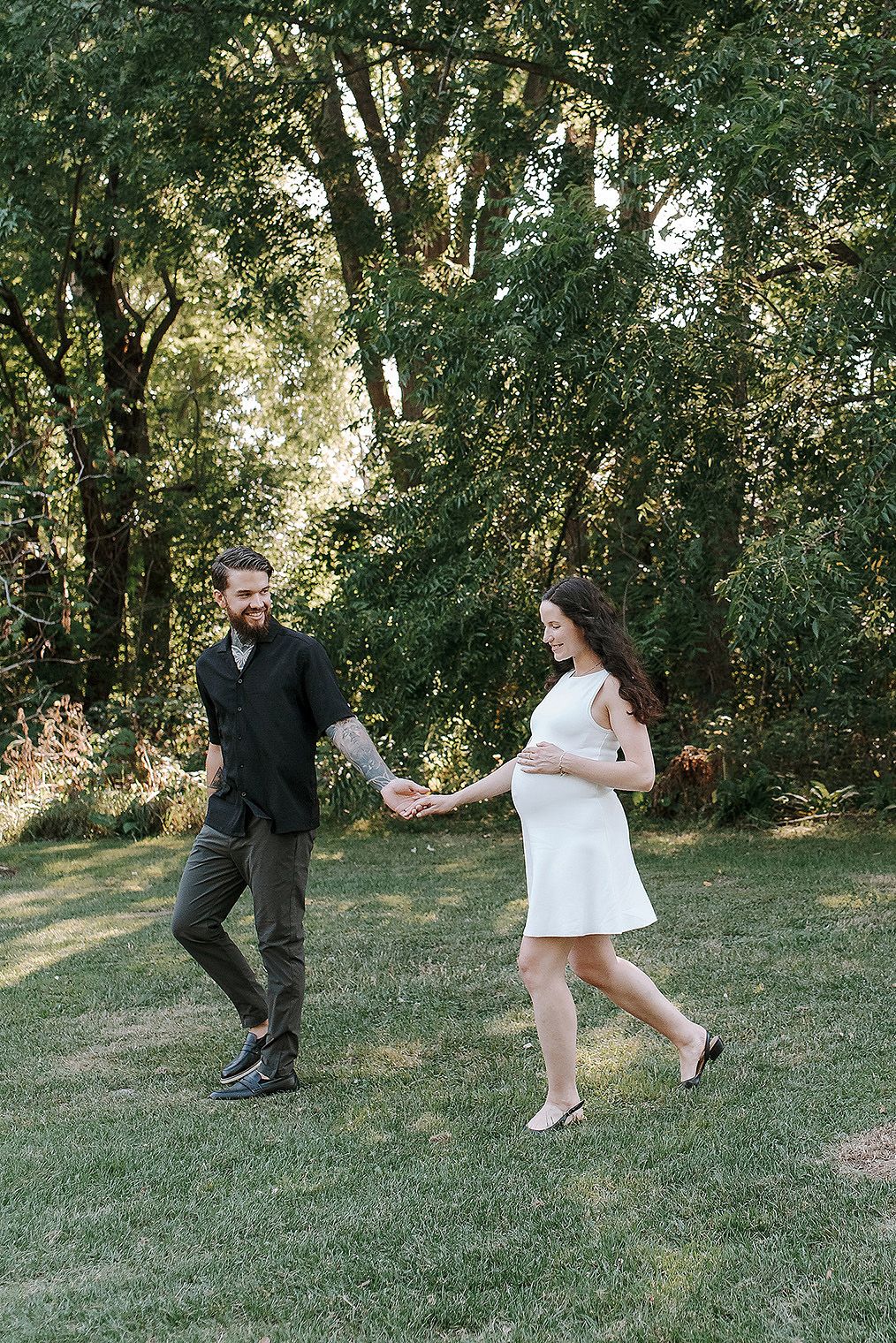 A couple dances together on a grassy lawn surrounded by lush green trees on a sunny summer day.