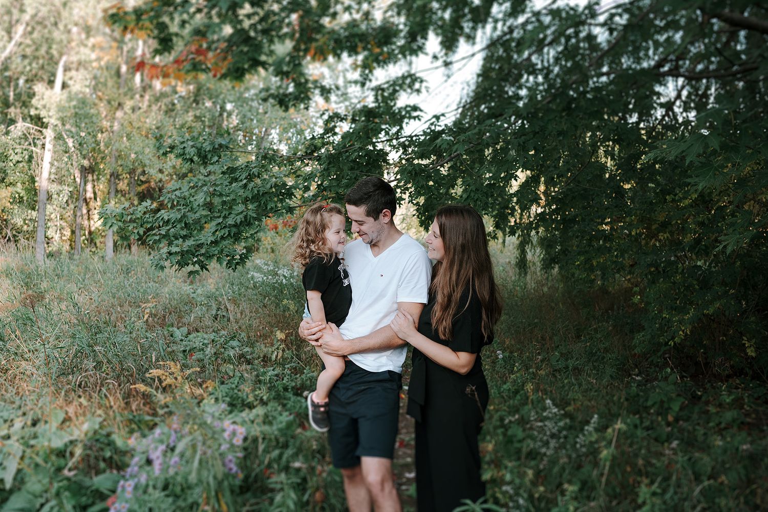 A couple stands together in a lush green outdoor setting during a summer photo session.