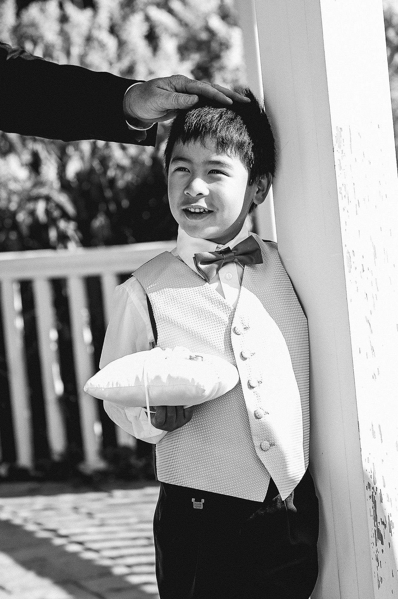 A black and white portrait of a young child in formal attire smiling while leaning against a white pillar outdoors.
