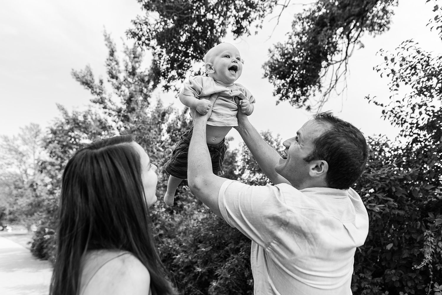 Black and white photo of parents lifting a joyful baby outdoors in a garden setting.
