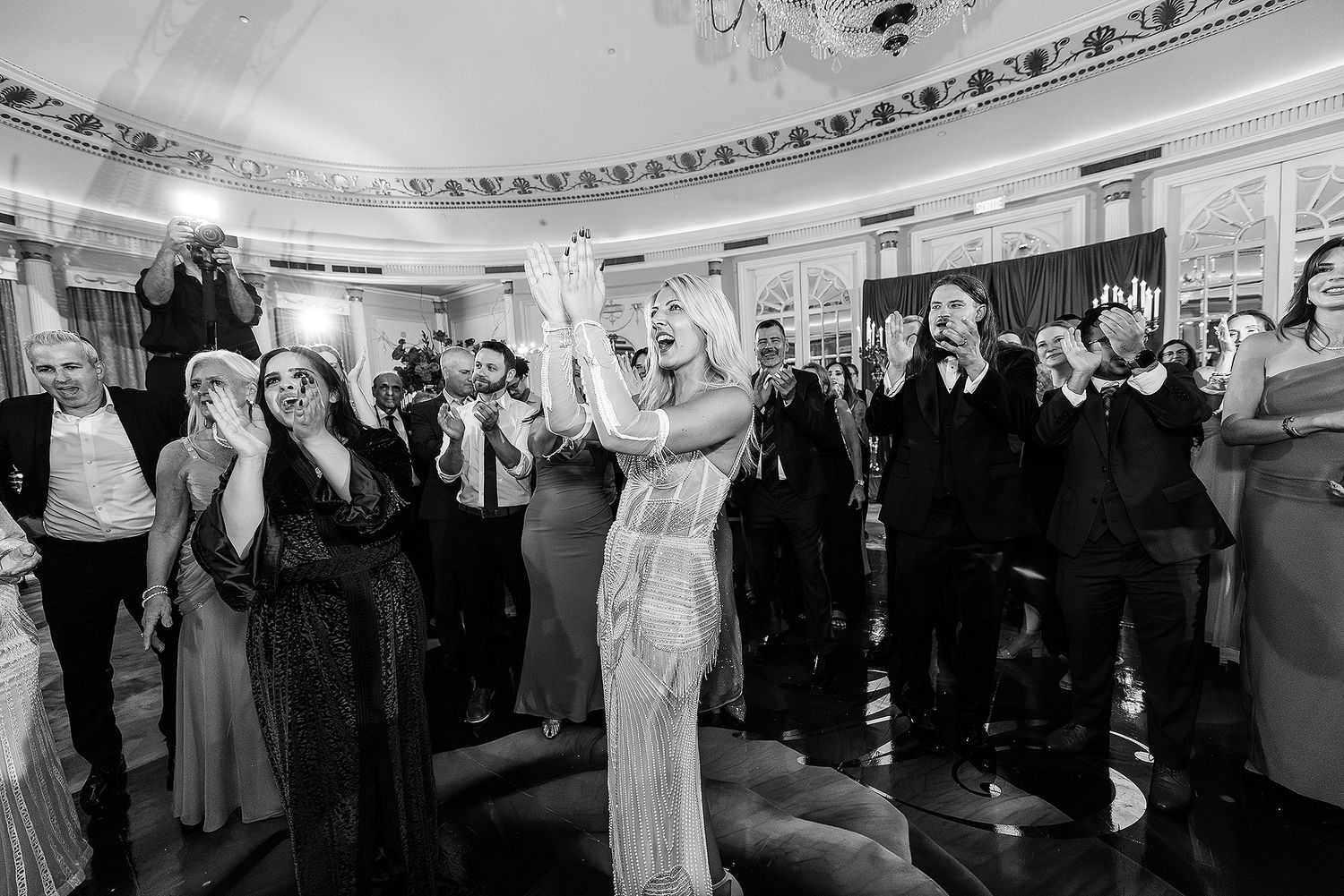 A wedding reception scene with guests dancing and celebrating in an elegant ballroom with ornate ceiling details.