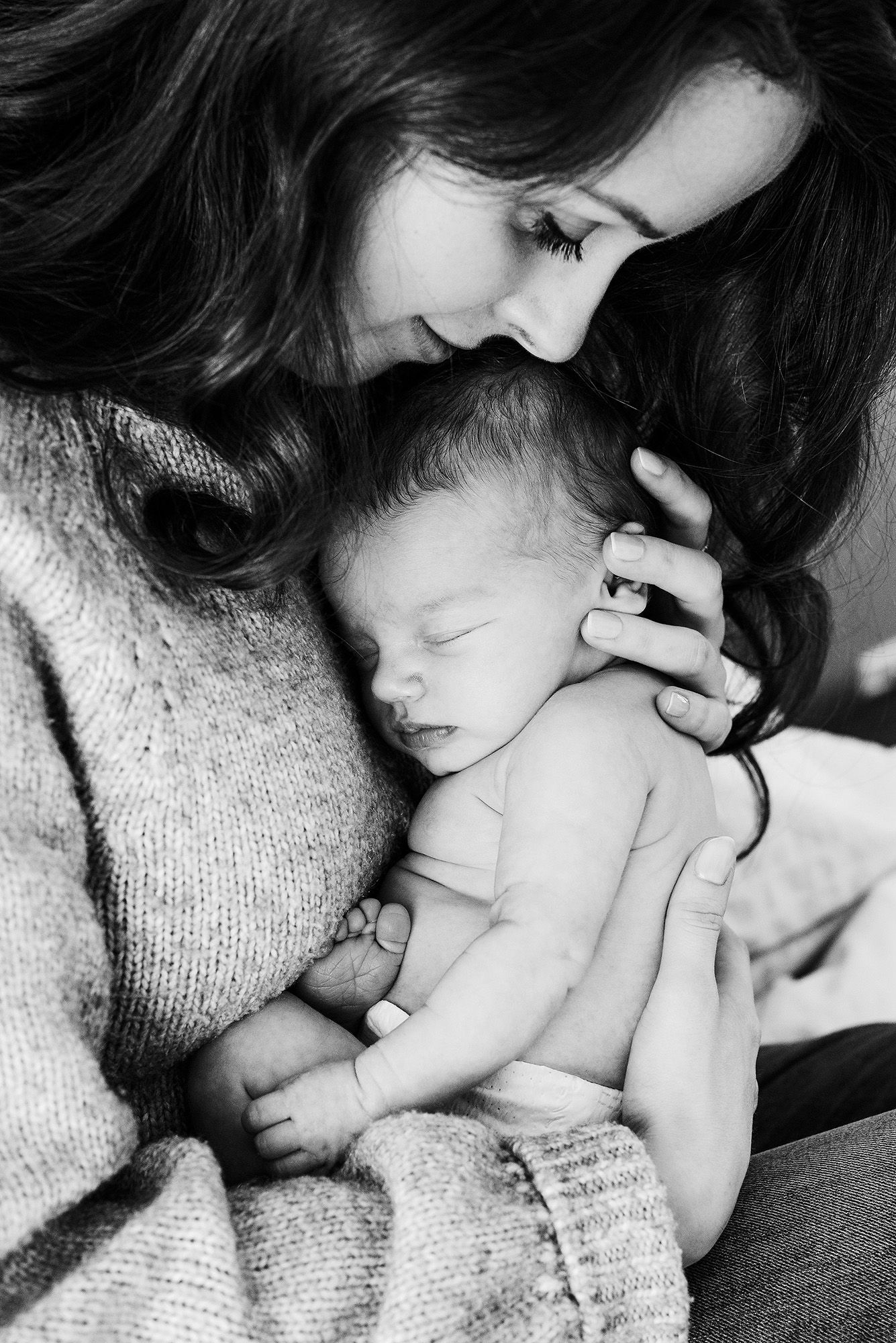 Intimate black and white portrait of parent snuggling with newborn on textured blanket.