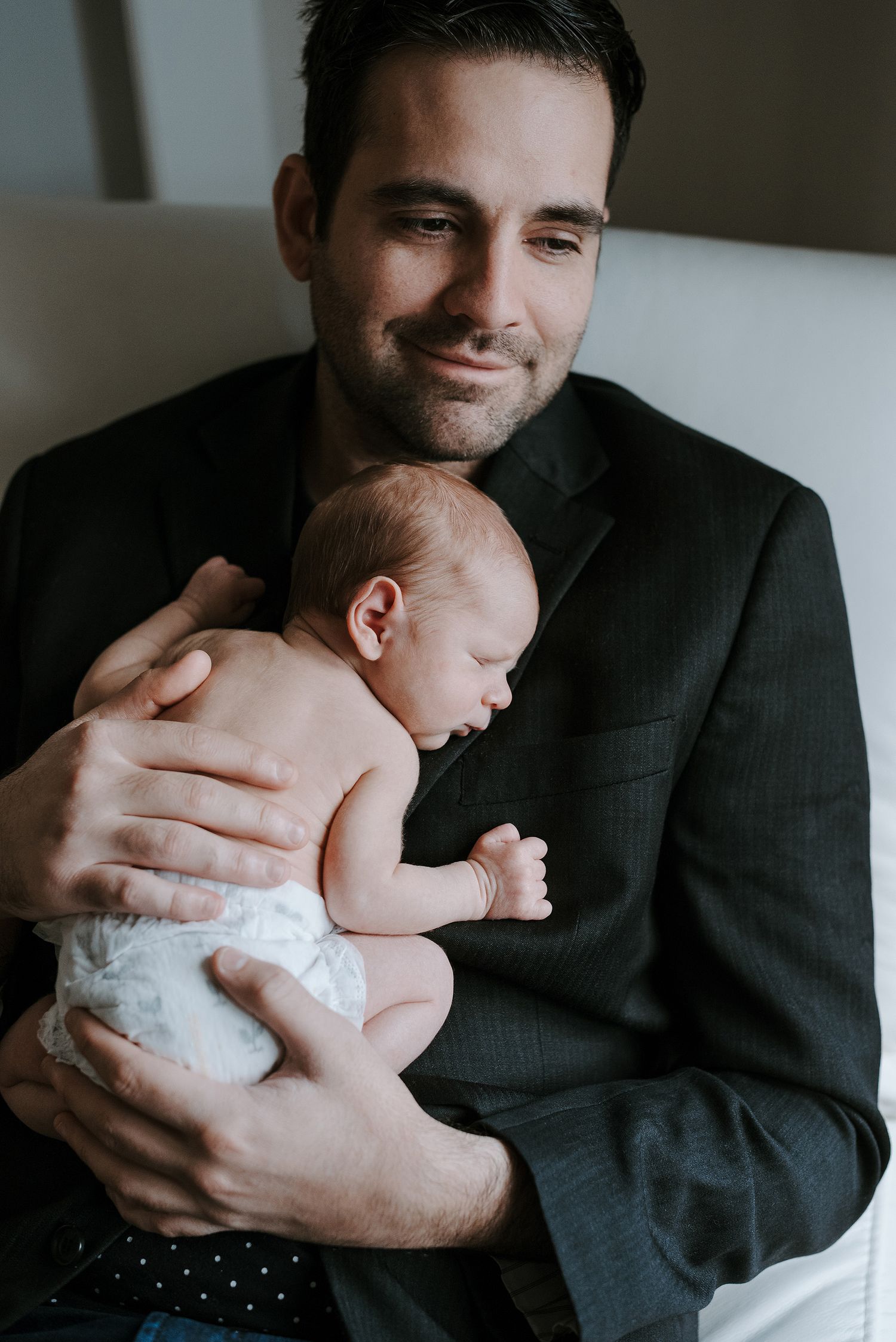 A newborn baby rests peacefully against a dark suit in an indoor setting.
