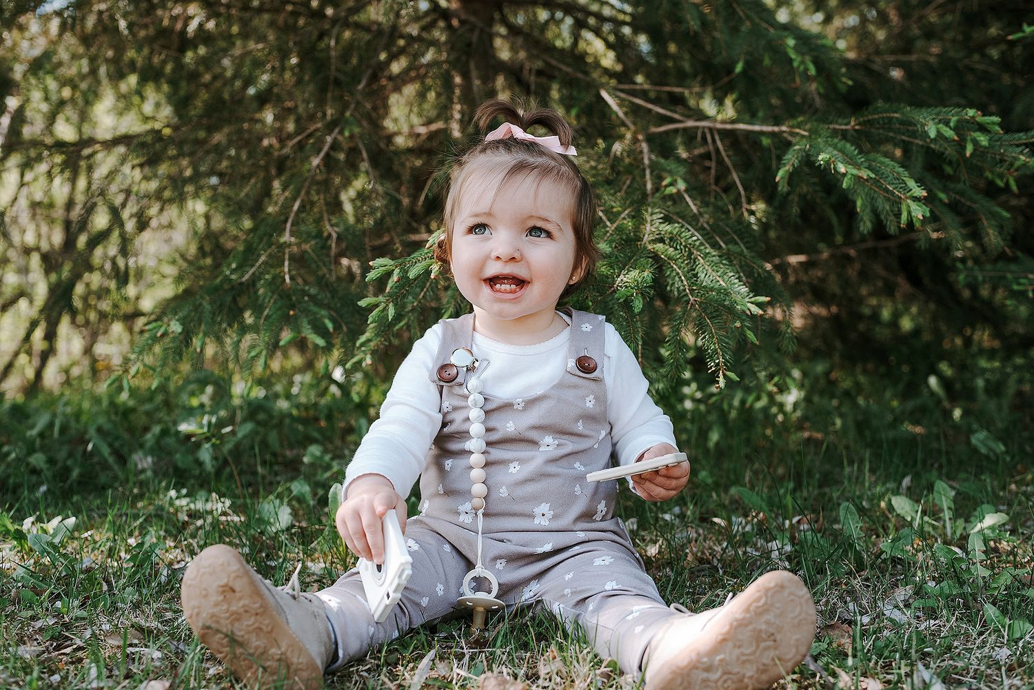 A toddler in gray overalls sits on grass with evergreen trees in the background.