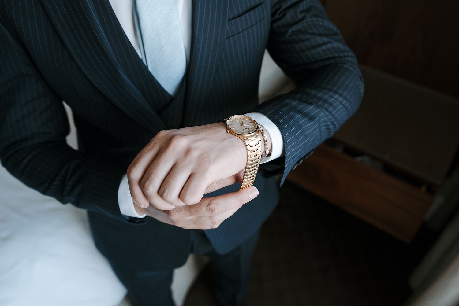 A person in a dark suit adjusts their watch while sitting in a dimly lit room.