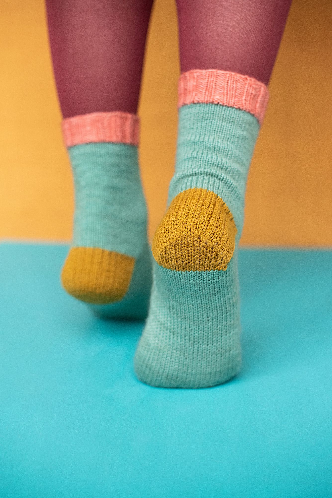 Close-up view of mint green and mustard yellow color-blocked knitted socks on feet against turquoise background.