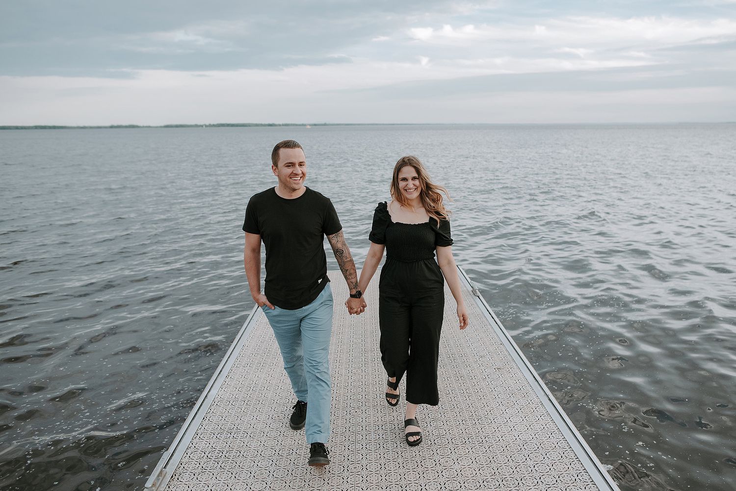 A couple walking hand in hand on a pier overlooking calm waters.