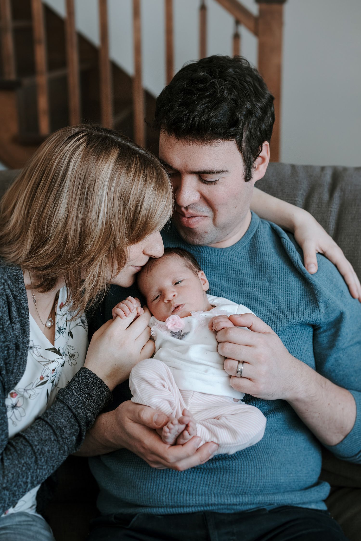 Parents cuddle with their newborn baby on a grey couch in a cozy living room setting.