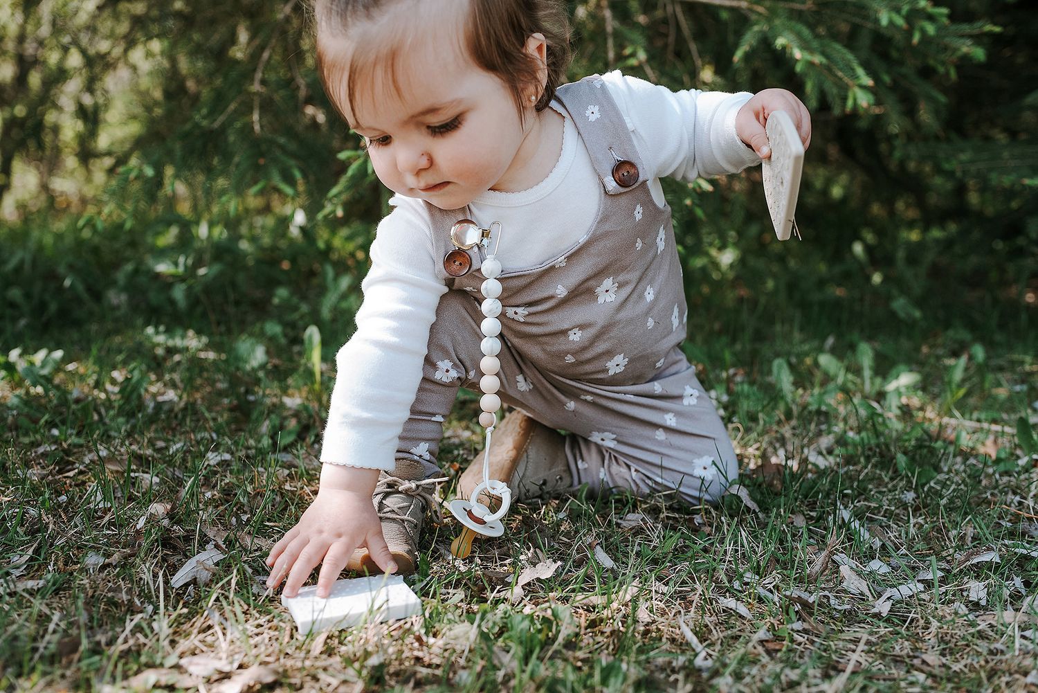 A small child in gray overalls bends down to explore something on the ground in a wooded area.