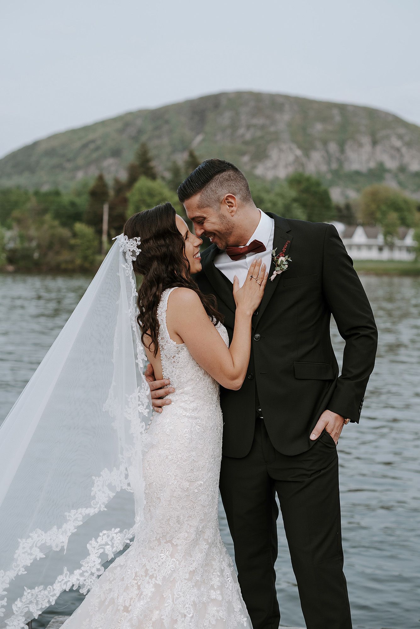 Romantic lakeside wedding photoshoot with bride in lace gown and groom in black suit against mountain backdrop.