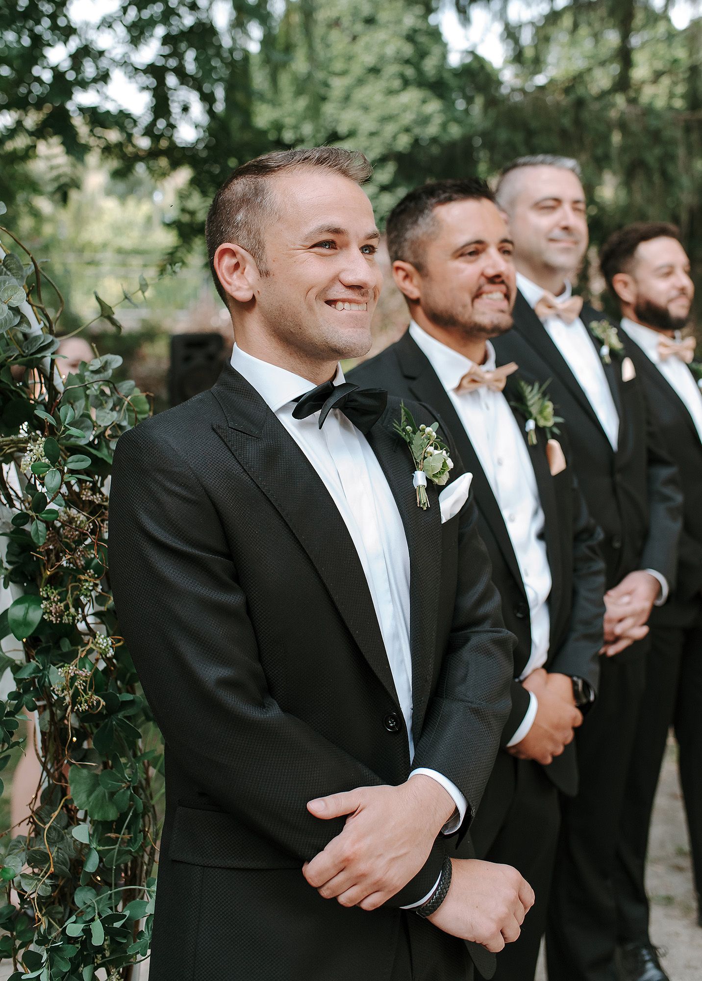 Groomsmen in classic black tuxedos and bow ties share a candid moment together outdoors at a formal wedding.