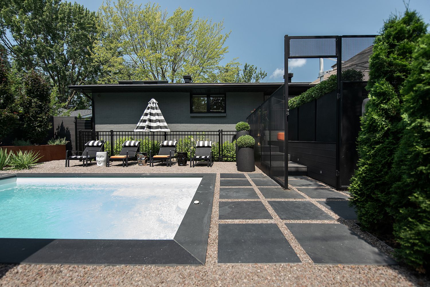 Modern backyard with a sleek black pool house, swimming pool, and geometric concrete patio design surrounded by greenery.