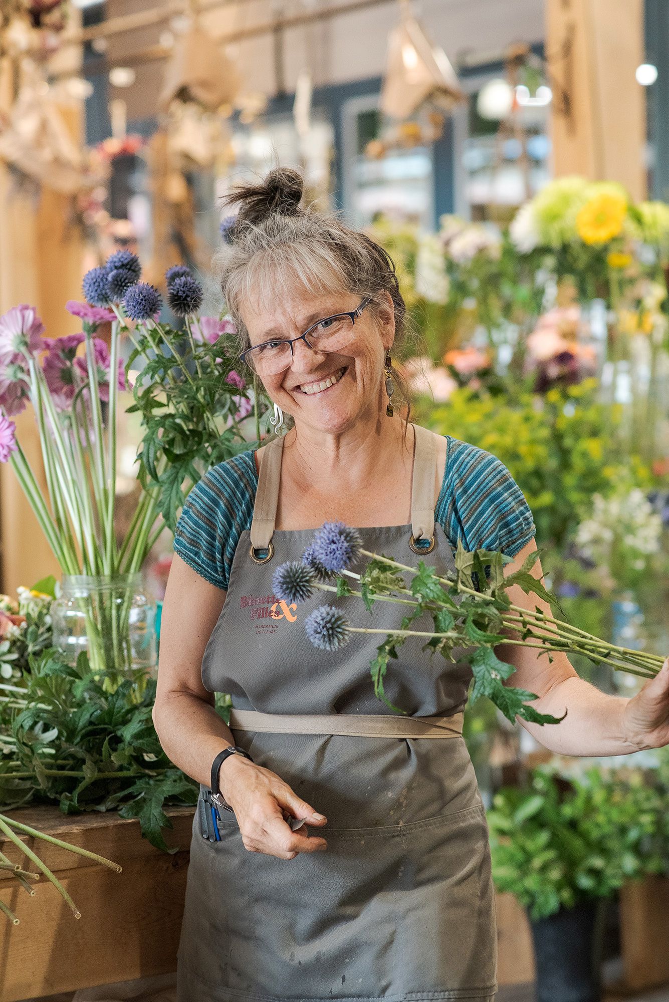 Florist in gray apron holding fresh purple thistle flowers in a bright flower shop display.