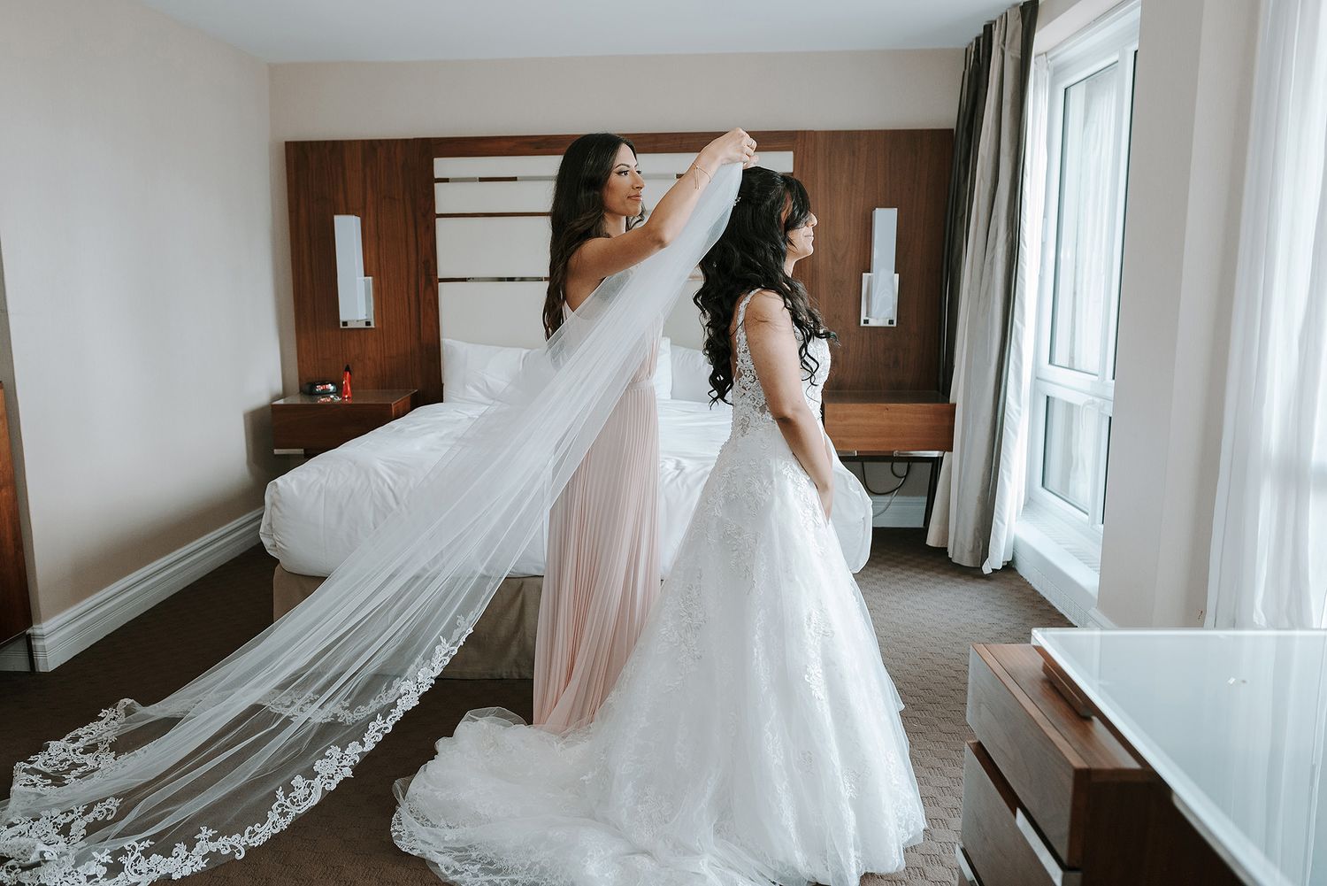 A flowing white wedding gown and veil being adjusted in an elegant hotel room with wood paneled walls.