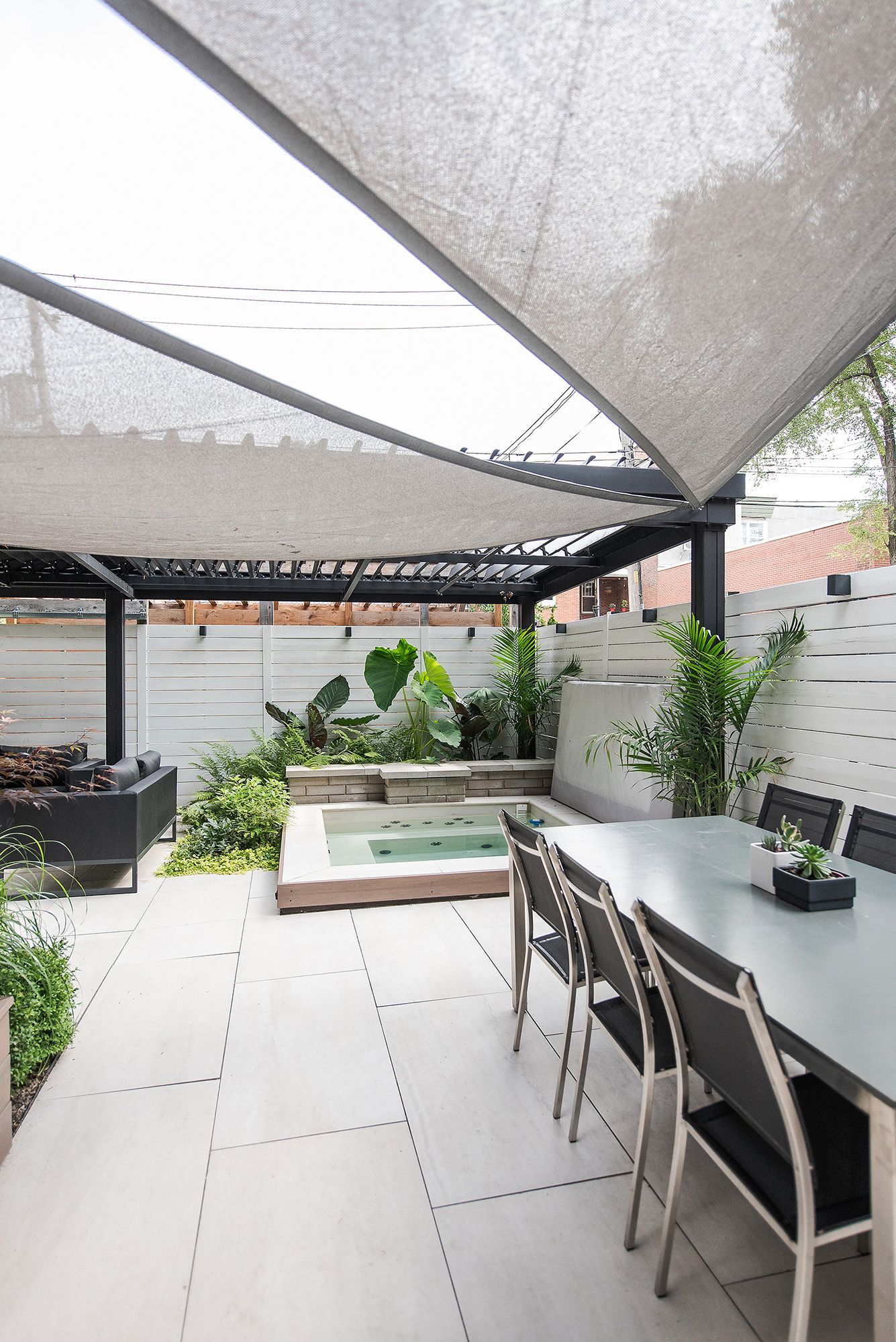 Modern outdoor patio with covered dining area, gray tile flooring, and tropical plants features a built-in spa and shade sails.