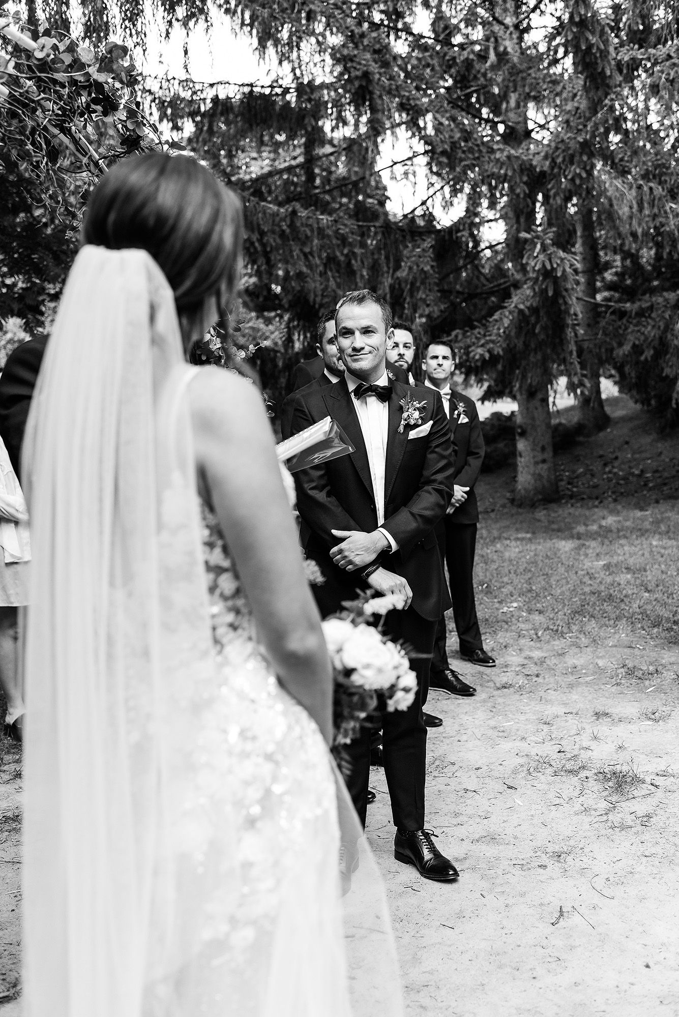 Black and white wedding photo showing first look moment between couple on a tree-lined path during outdoor ceremony.
