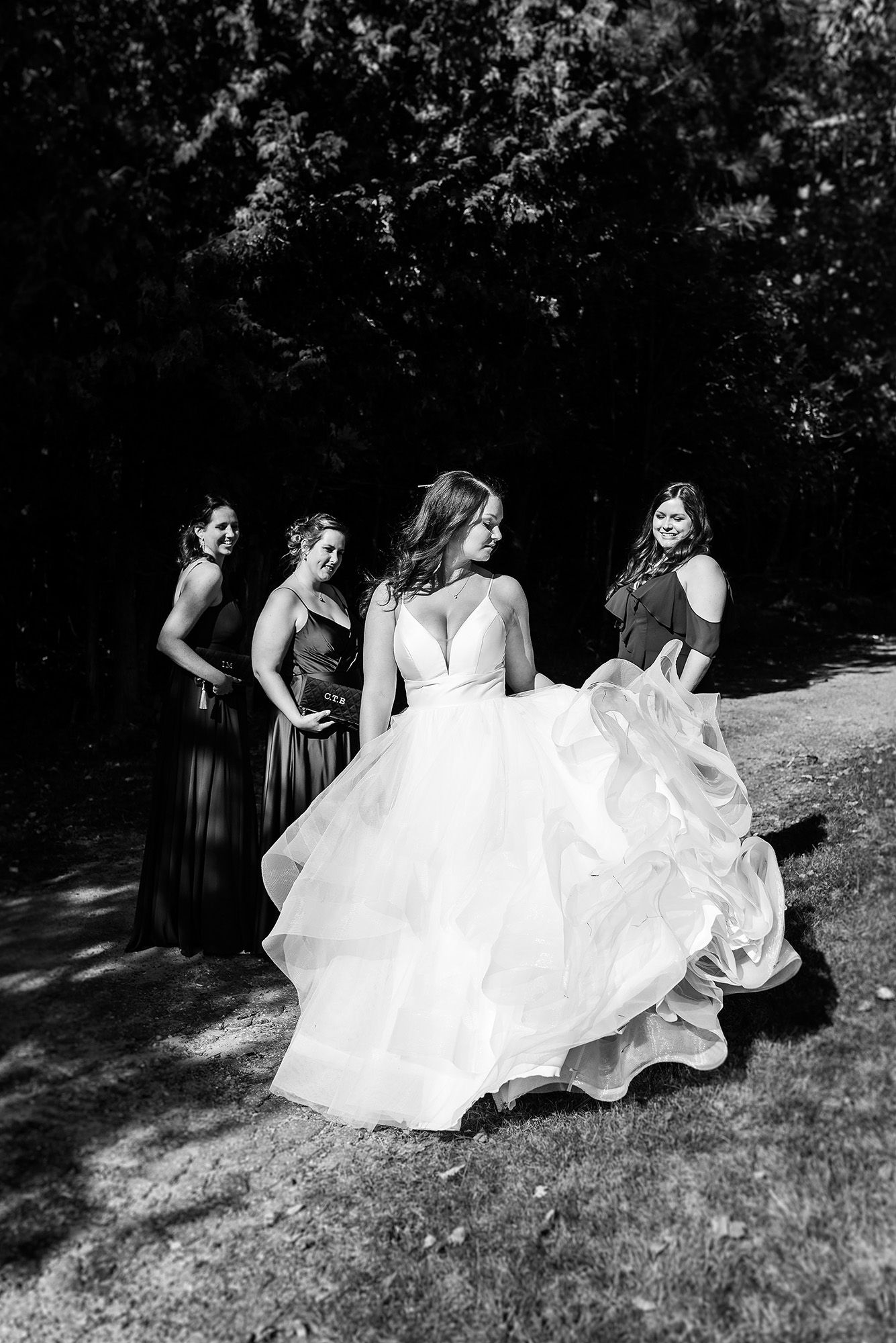 A bride in a flowing white gown twirls outdoors while bridesmaids in dark dresses look on during a sunny wedding day.
