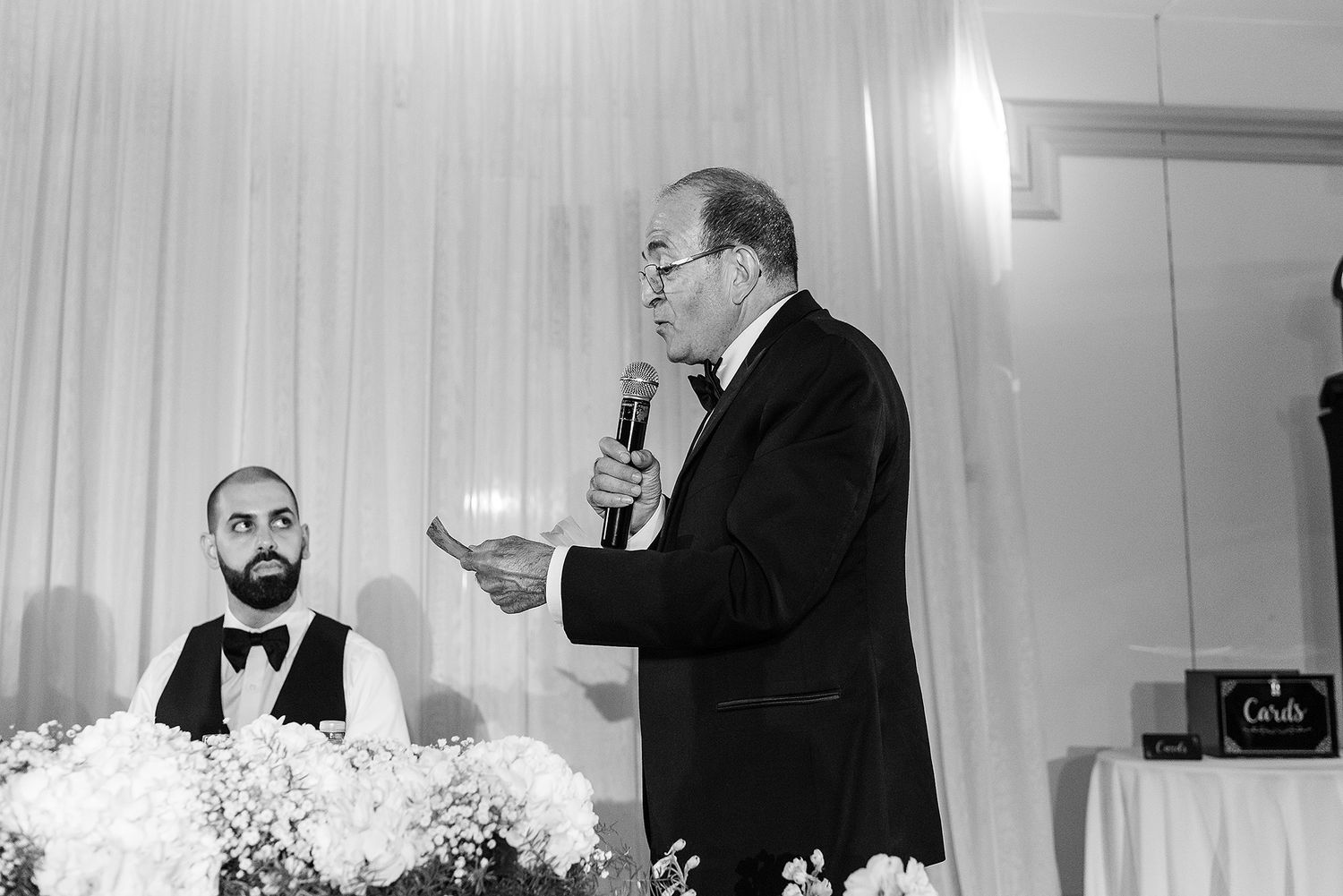 A person in a tuxedo speaks into a microphone at a formal event with white floral decorations on the table.