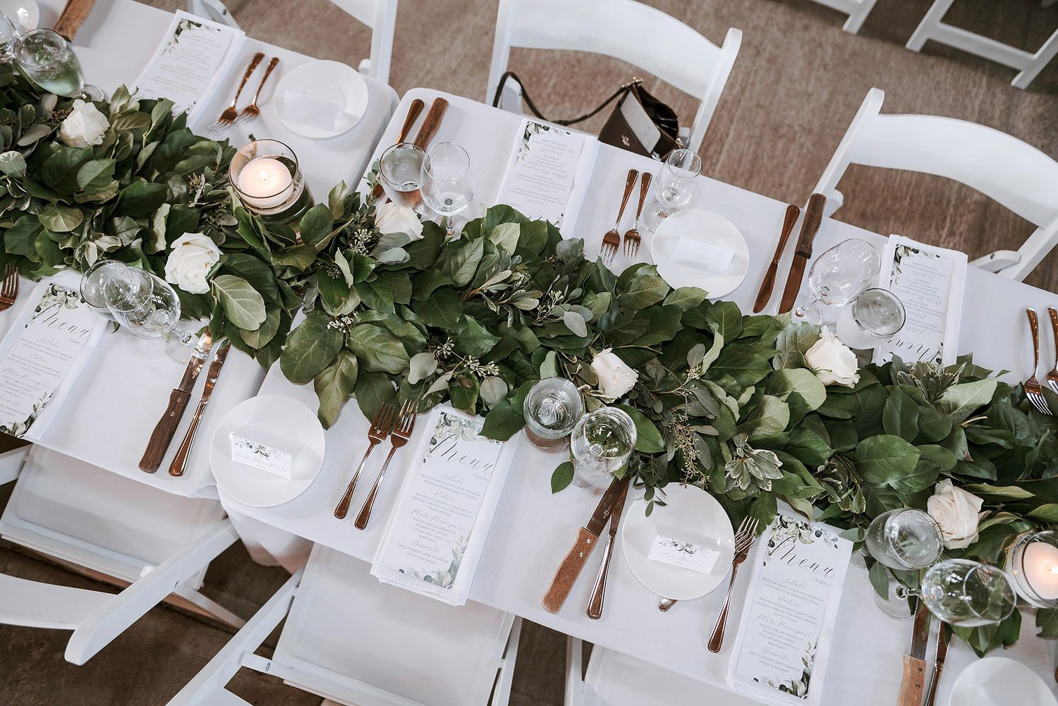 White wooden chairs line a rustic table decorated with lush greenery garland and place settings for an elegant event.