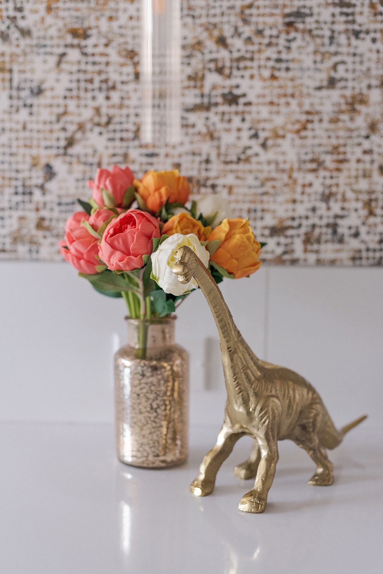 Gold dinosaur figurine next to pink and orange roses in a metallic vase against a textured wall.