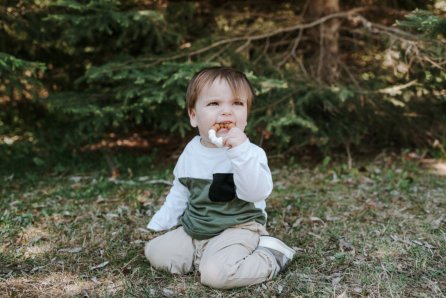 Baby in white and gray outfit sitting on grass in a natural outdoor setting.