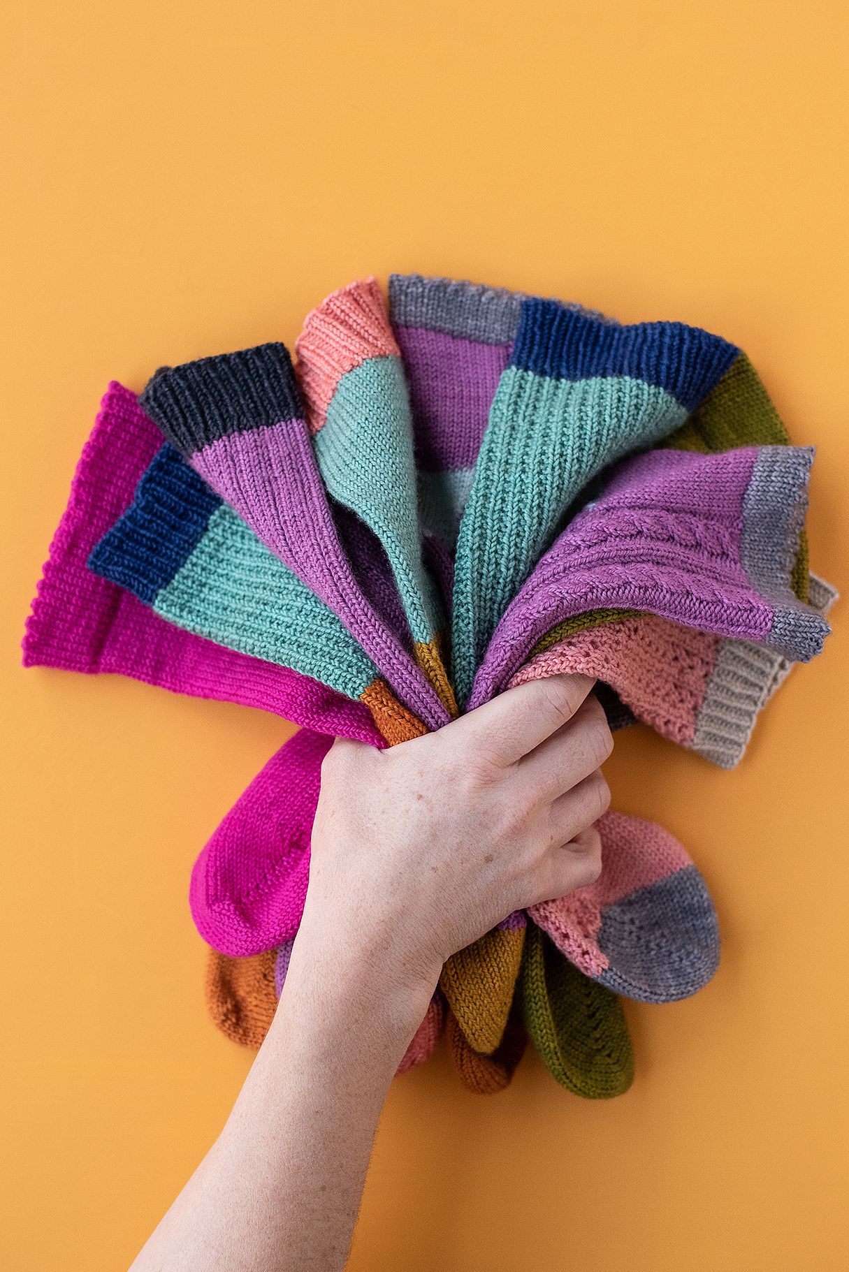 Hand arranging multiple colorful knitted socks in a fan pattern on orange background.