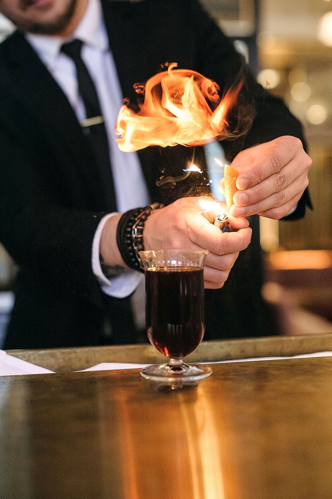 Bartender lighting cocktails on fire in dramatic fashion across multiple glasses at an upscale bar counter.