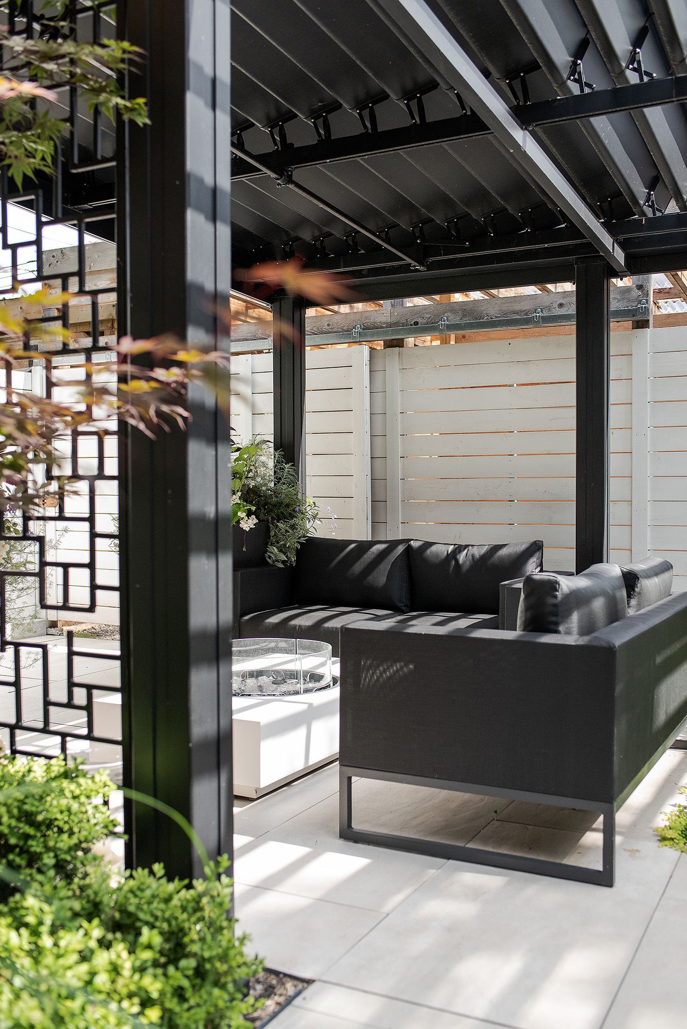 Modern outdoor pergola with black beams and hanging swing seating against a white privacy wall.