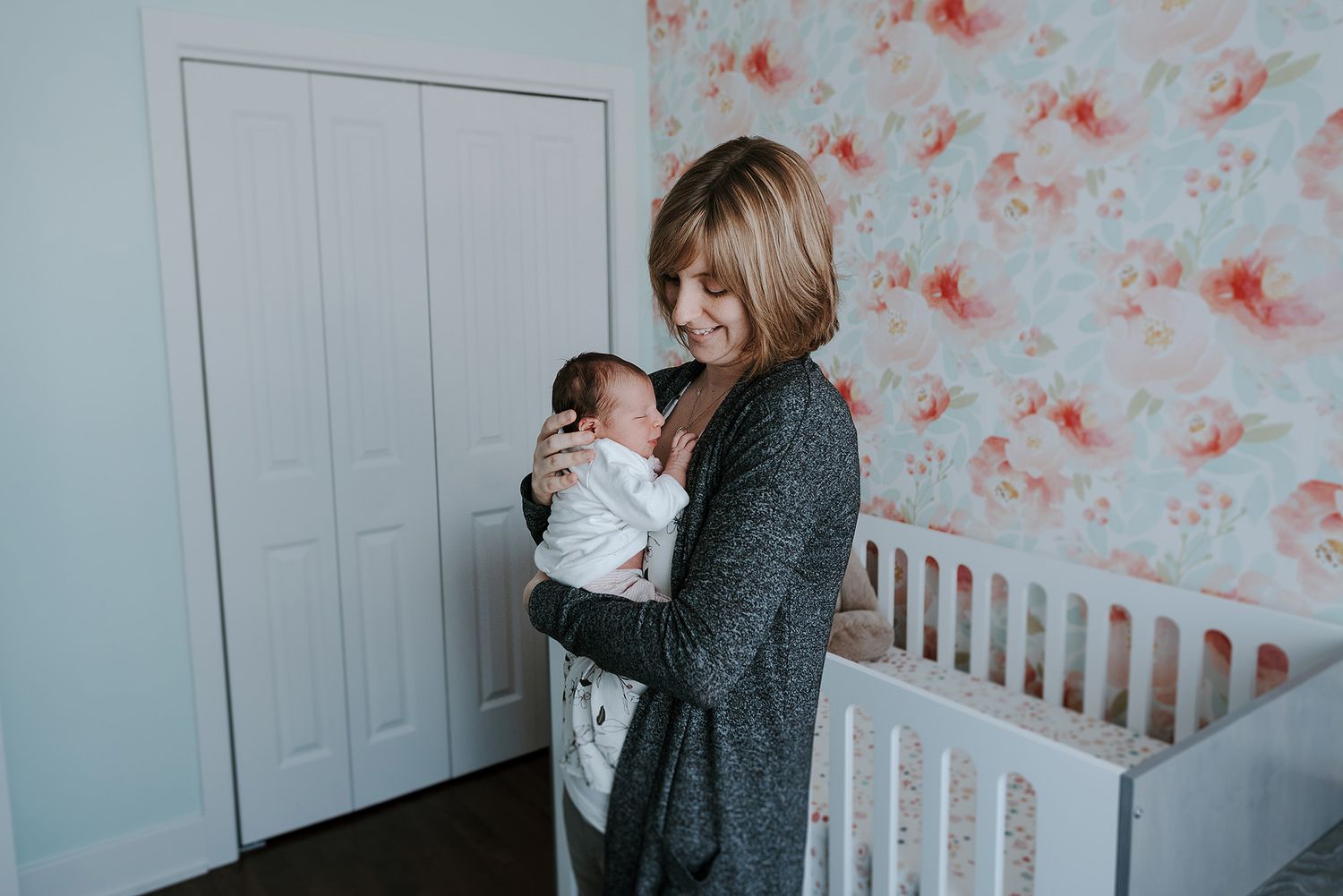 A nursery scene with floral wallpaper showing a parent holding a swaddled newborn.