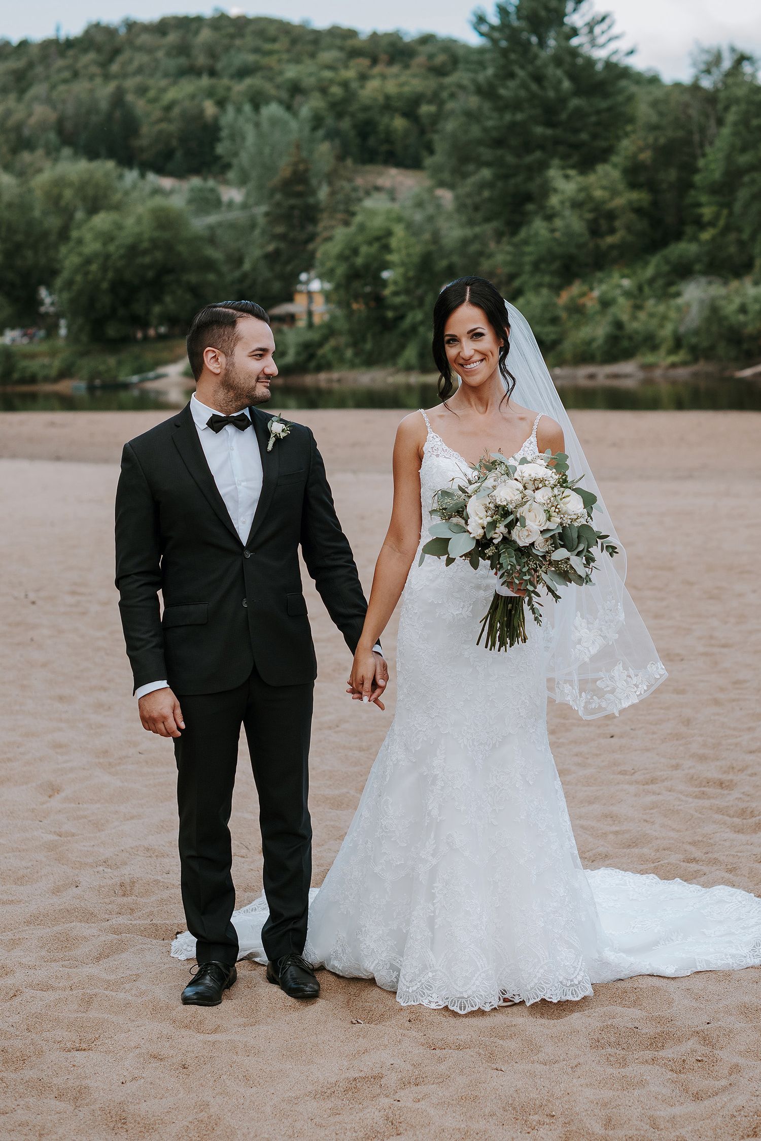 A couple in wedding attire walks along a sandy beach with trees in the background during their romantic photo session.