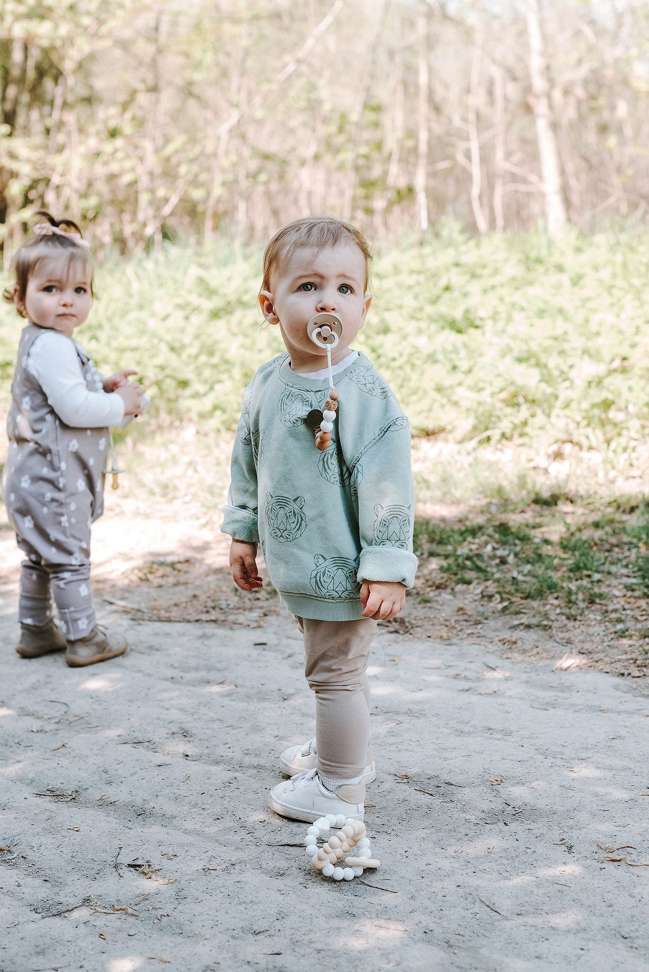 Toddler in mint green cardigan walking outdoors while holding a pacifier.