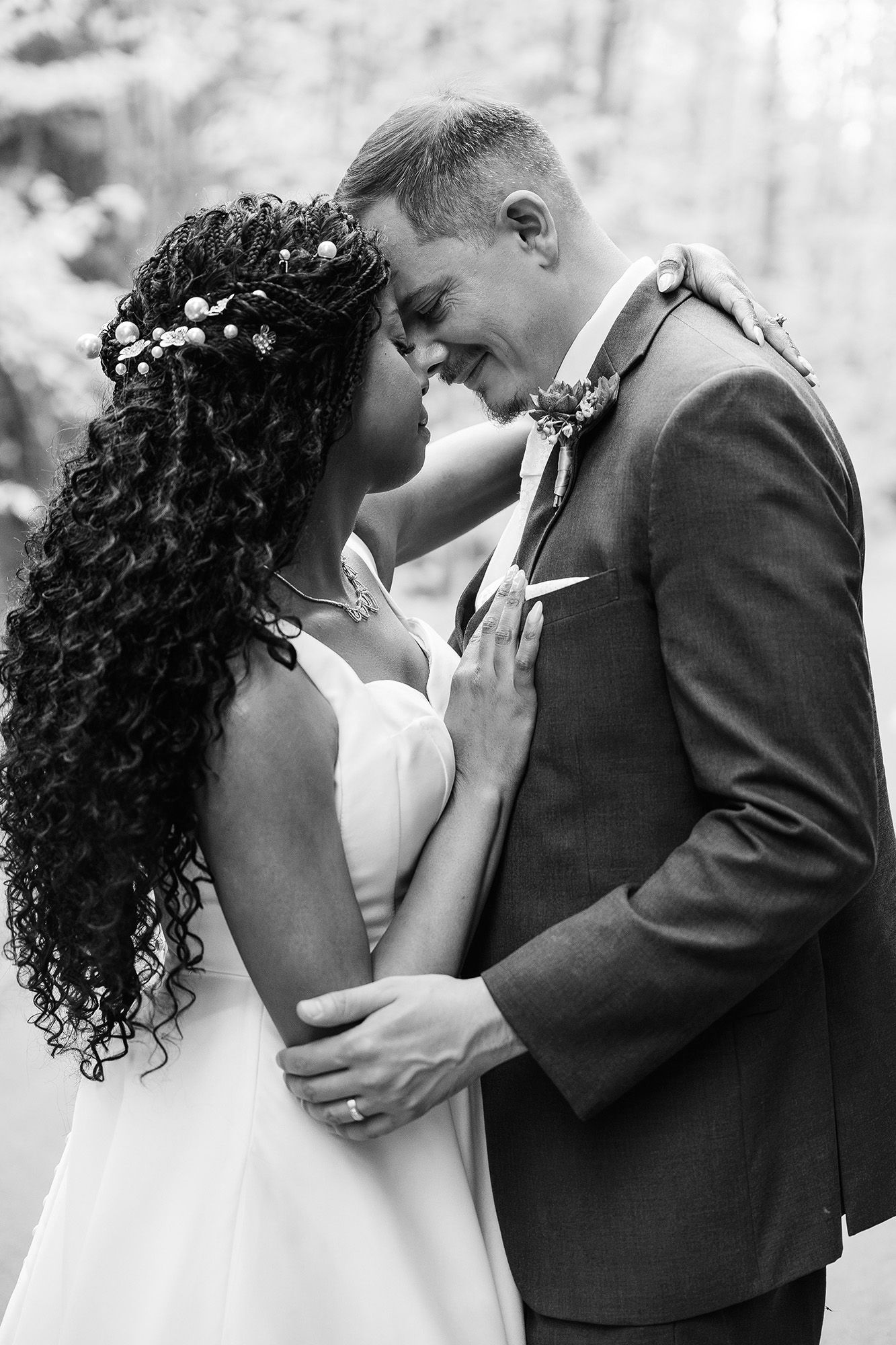 Black and white photo of a smiling interracial couple in wedding attire embracing forehead-to-forehead outdoors.