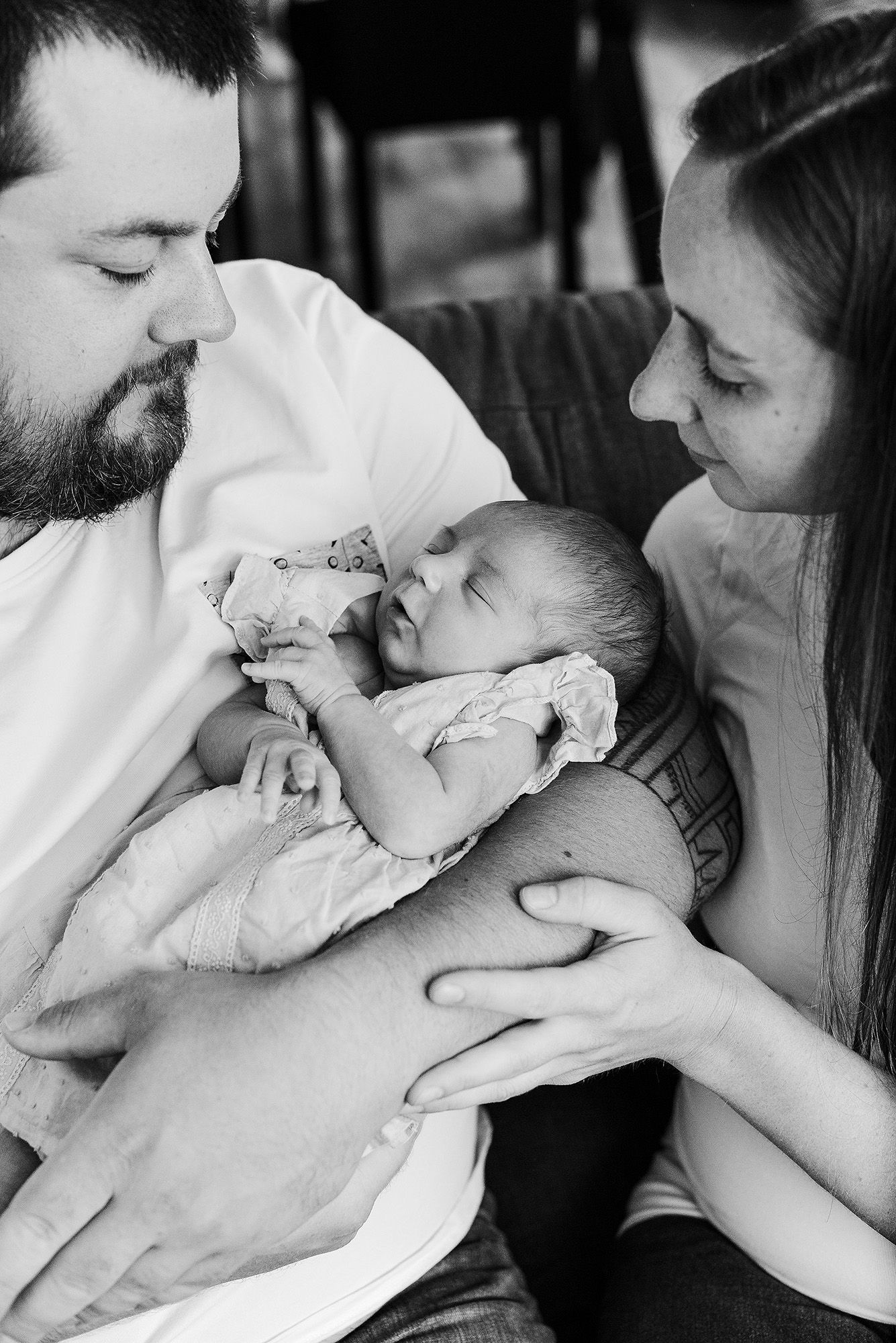 An intimate black and white portrait of parents gazing at their sleeping newborn on a couch.