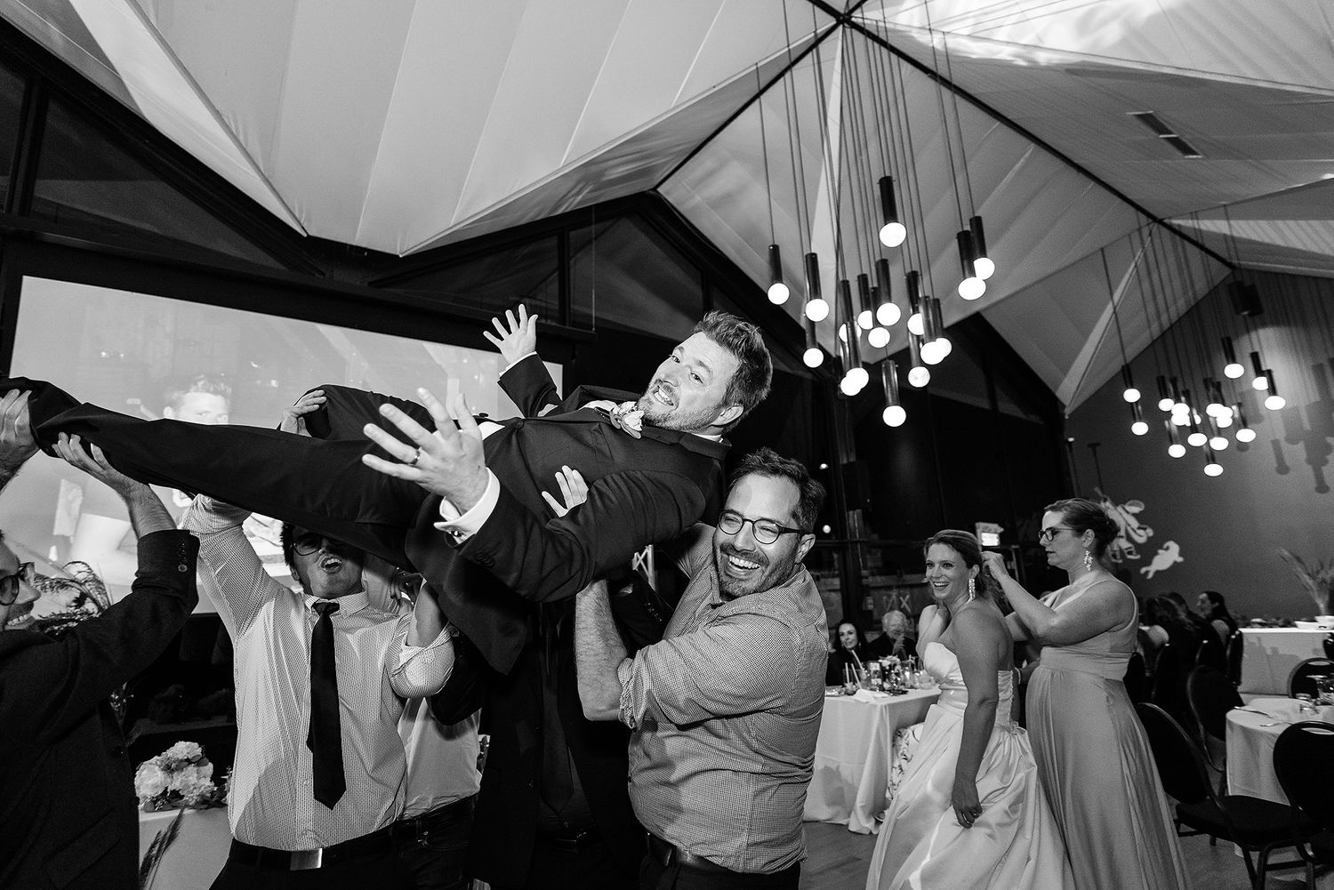 Black and white photo series showing wedding reception guests dancing and celebrating under a tent with hanging lights.