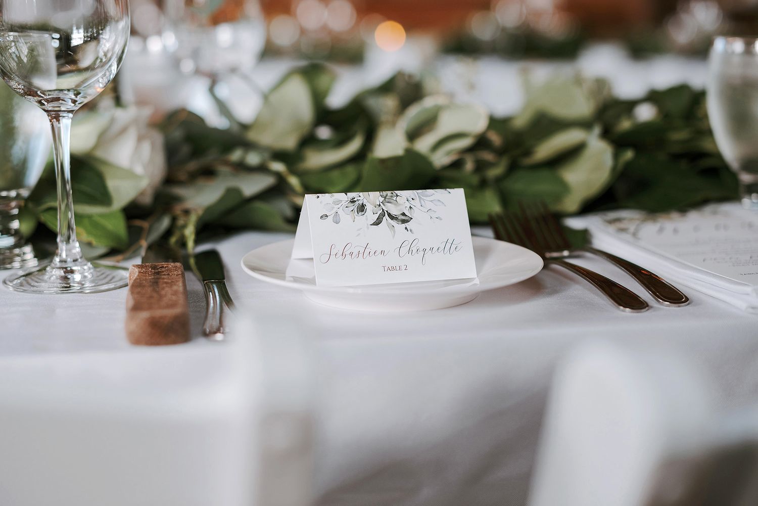 Elegant wedding reception table setting with white linens, wine glasses, and delicate greenery decorations.