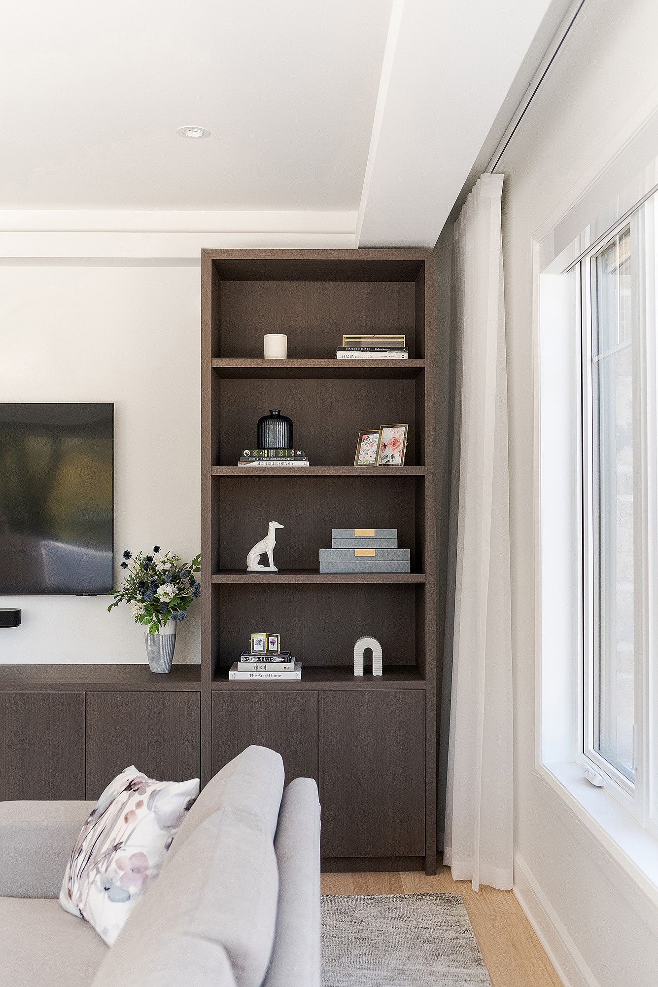 Modern living room interior with dark wood built-in shelving, white walls, and natural light from large windows.