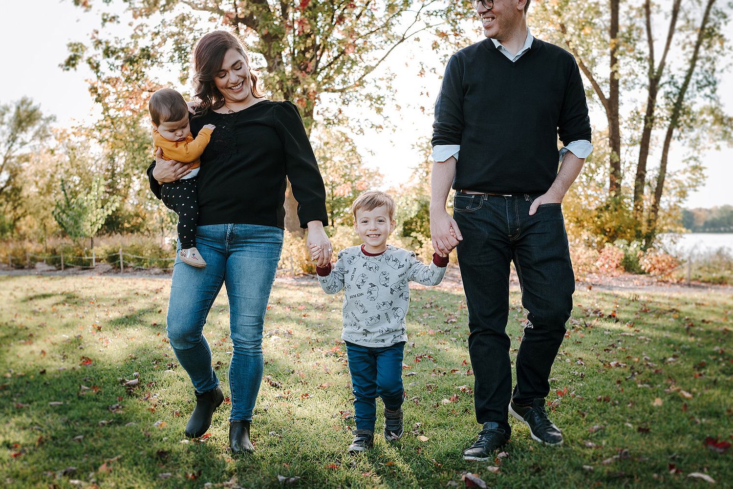 A family walks together and plays in a scenic autumn setting with fallen leaves and tall trees.