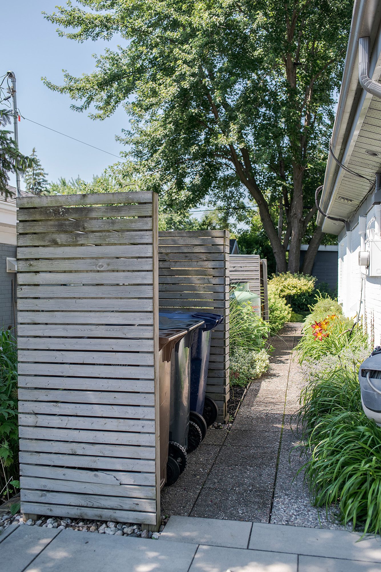 Modern wooden privacy screen conceals garbage bins along a narrow side yard path with landscaping and trees.