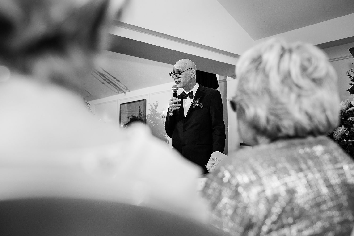 Black and white candid wedding photo showing someone adjusting their tie in a mirror during preparations.
