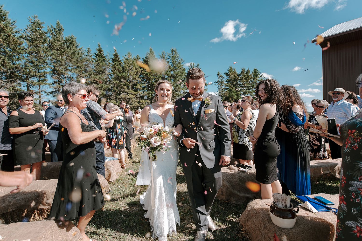 Wedding guests celebrate outdoors with raised hands against a backdrop of evergreen trees and blue sky.