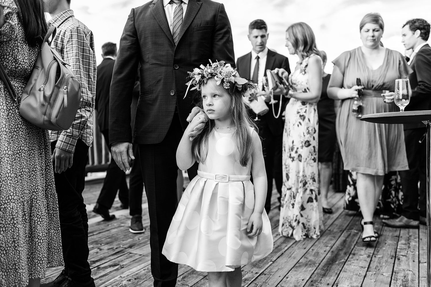 A young flower girl in a white dress and floral crown stands on a wooden deck during a wedding celebration.