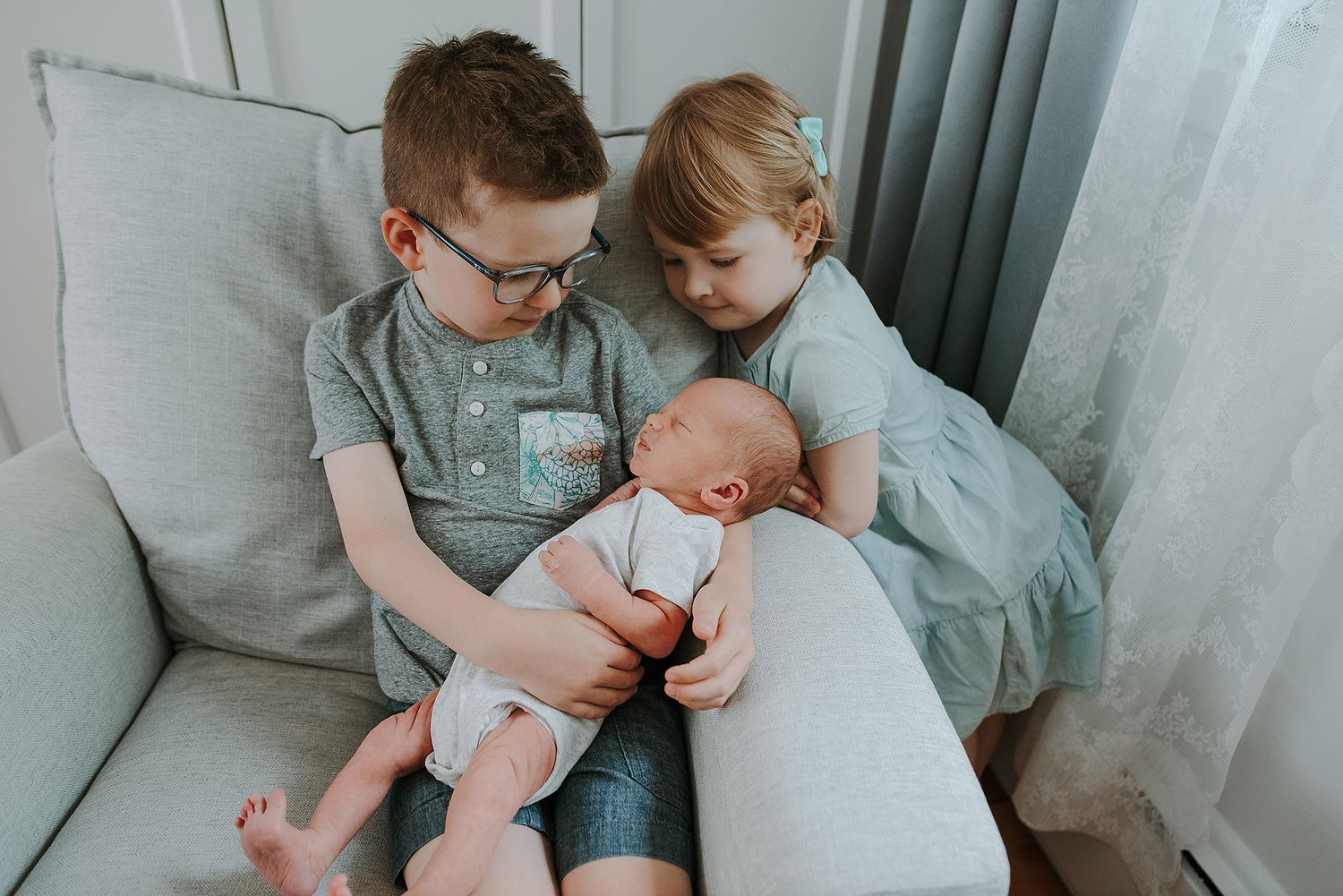 Two children and a baby share a tender moment on a grey couch.